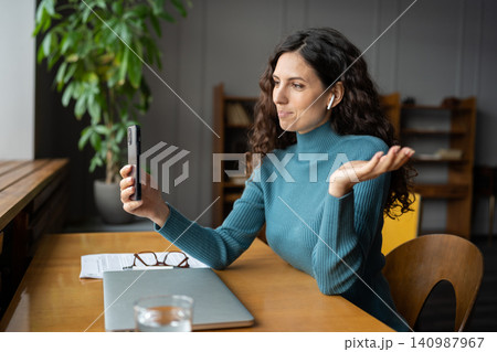Smiling young businesswoman in earbuds sitting at office desk talking by video call on smartphone 140987967