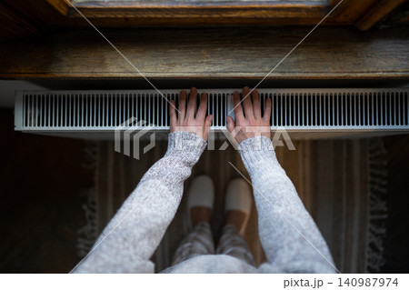 Woman in woolen sweater tries to catch stream of warm air from central heater touching device 140987974