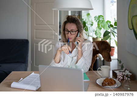 Employee woman sitting on chair with dog at home, taking on mobile phone, remote working at laptop 140987976