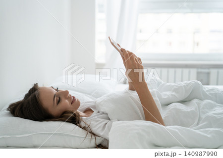 Smiling young woman using mobile smart phone, lying and relaxing in white comfortable bed at home 140987990
