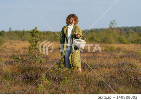 Nature autumn activities. Young woman searching cranberries on swamp holding basket happy smiling Nature autumn activities. Young woman searching cranberries on swamp holding basket happy smiling 140988002