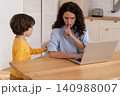 Mom sits by table at home office during lockdown, working on laptop, kid distracts and making noise 140988007