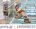 Focused female student enjoys reading interesting book with orange cover sitting on glass stairs 140988010
