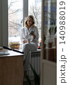 Woman sitting on windowsill using mobile smart phone holding mug, drinking coffee in morning at home 140988019