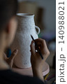 Female artisan paints text on white ceramic vase with marker on blurred background in workshop 140988021