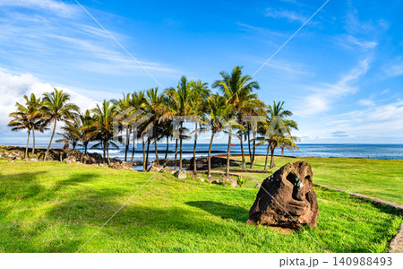 Ancient Moai stone head on green grass surrounded by palm trees and the ocean under a blue sky on Easter Island, Chile Ancient Moai stone head on green grass surrounded by palm trees and the ocean under a blue sky on Easter Island, Chile 140988493