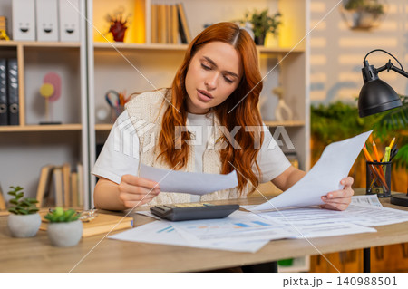 Young woman on home table paying bills with calculator and checking receipts during home budget task Young woman on home table paying bills with calculator and checking receipts during home budget task 140988501