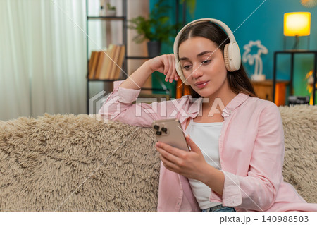 Young woman listening to podcast on sofa wearing headphones nodding while writing insightful notes Young woman listening to podcast on sofa wearing headphones nodding while writing insightful notes 140988503