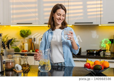 Young woman pouring cold lemon water in home kitchen to quench thirst for diet hydration fit routine Young woman pouring cold lemon water in home kitchen to quench thirst for diet hydration fit routine 140988586