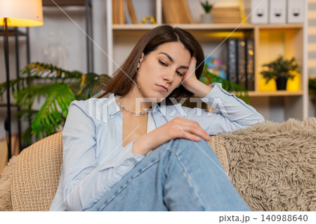 Young woman on home sofa feeling sad and worried thinking deeply and shaking head in concern indoors Young woman on home sofa feeling sad and worried thinking deeply and shaking head in concern indoors 140988640