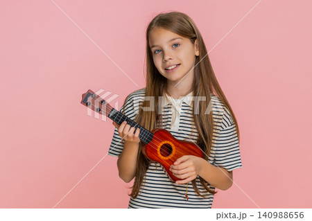 Little child girl kid playing toy ukulele smiling while enjoying imaginary concert performance Little child girl kid playing toy ukulele smiling while enjoying imaginary concert performance 140988656
