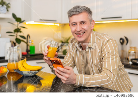 Middle-aged man using smartphone while drinking orange juice in kitchen for diet rest and fun time Middle-aged man using smartphone while drinking orange juice in kitchen for diet rest and fun time 140988686
