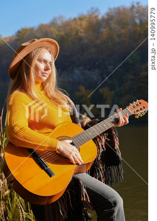Beautiful blonde girl playing acoustic guitar and singing relaxing enjoying outdoors on sunny autumn day. Hippie woman in boho style learning and practicing playing musical instrument. Fall leaves 140995779