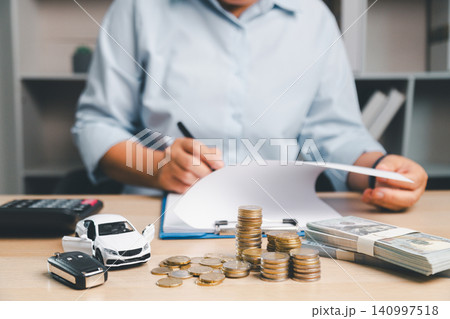 Business person reviewing car finance documents with white toy car, car key, cash, and coin stacks on desk, representing vehicle loan approval, auto insurance, and purchase cost planning. Business person reviewing car finance documents with white toy car, car key, cash, and coin stacks on desk, representing vehicle loan approval, auto insurance, and purchase cost planning. 140997518