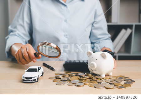 Person using a magnifying glass to inspect car finance and savings items with piggy bank, toy car, coins, and calculator, representing vehicle insurance review and loan analysis. Person using a magnifying glass to inspect car finance and savings items with piggy bank, toy car, coins, and calculator, representing vehicle insurance review and loan analysis. 140997520