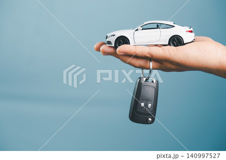 White toy car with car key hanging from a hand, showing auto insurance, car rental, vehicle leasing, ownership transfer, car loan, and transportation service concept. White toy car with car key hanging from a hand, showing auto insurance, car rental, vehicle leasing, ownership transfer, car loan, and transportation service concept. 140997527