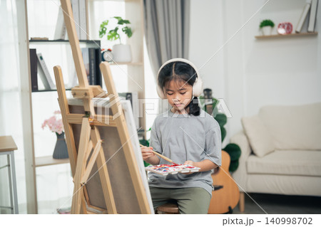 Young girl wearing headphones painting on canvas using brush and palette in cozy bright living room with artistic decor and comfortable furniture 140998702