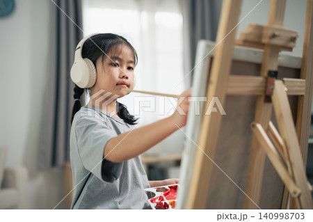Young Asian girl wearing white headphones painting on easel with focused expression in cozy indoor room with soft natural light Young Asian girl wearing white headphones painting on easel with focused expression in cozy indoor room with soft natural light 140998703