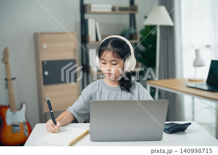 Young girl wearing headphones studying at home using laptop and writing in notebook focused on online learning and education 140998715