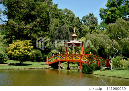 Traditional red Japanese bridge crossing a peaceful pond in a beautiful botanical garden. Scenic landscape with lush trees, calm water and elegant oriental architecture on a sunny day. 141000080
