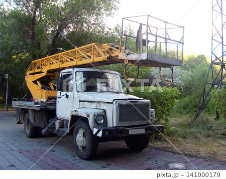 An old cherry picker truck with a white cabin and a yellow telescopic boom lift parked on a paved road. The cherry picker vehicle is surrounded by green trees and bushes on a bright summer day An old cherry picker truck with a white cabin and a yellow telescopic boom lift parked on a paved road. The cherry picker vehicle is surrounded by green trees and bushes on a bright summer day 141000179