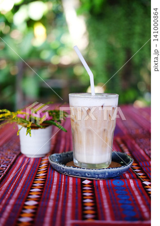 Fresh a clear glass of iced latte coffee with milk foam with blooming pink flowers in a glass vase on pink tablecloth with a Thai pattern on wooden table in resort of Thailand Fresh a clear glass of iced latte coffee with milk foam with blooming pink flowers in a glass vase on pink tablecloth with a Thai pattern on wooden table in resort of Thailand 141000864