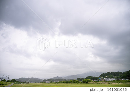 雨を降らせる大きな曇り空が広がる下で遠くに連なる山並みとのどかな集落や緑の広場の風景 141001086