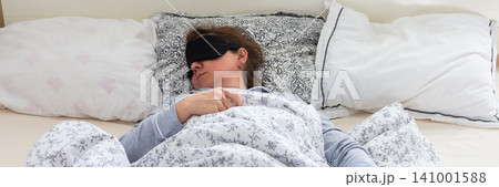 A 50-year-old woman is comfortably sleeping in her bed with a sleep mask on. She is wrapped in a soft blanket, resting peacefully in a serene bedroom setting, banner A 50-year-old woman is comfortably sleeping in her bed with a sleep mask on. She is wrapped in a soft blanket, resting peacefully in a serene bedroom setting, banner 141001588