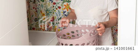 A woman stands in a laundry room holding a basket with clean clothes. She looks focused as she prepares to take out laundry from the washing machine. The room has bright floral wallpaper, banner 141001661