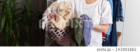 A person stands in a room holding a basket full of dirty clothes. Garments hang in the background. The setting is bright and casual, focusing on the laundry task ahead, banner 141001662