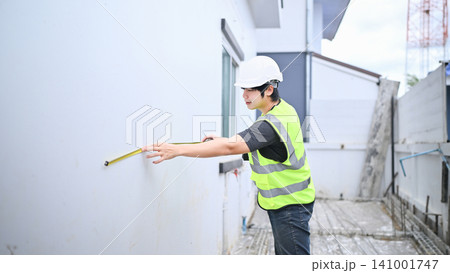 Asian Construction Engineer Inspector Measuring Wall with Tape Measure at Building Site 141001747
