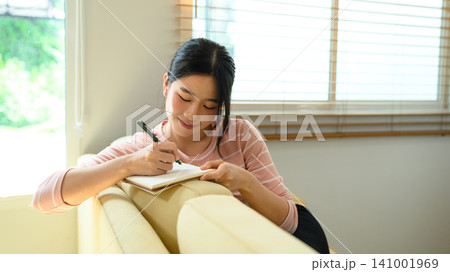 Young Asian Woman Smiling While Writing in Notebook on Sofa at Home 141001969