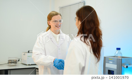 Two Female Scientists Shaking Hands in Laboratory Setting with Safety Equipment 141001970