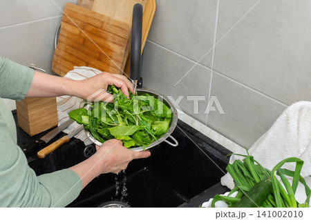 Hands are holding a colander filled with fresh organic local vegetables under running water in a kitchen sink. This is done to clean the produce before cooking Hands are holding a colander filled with fresh organic local vegetables under running water in a kitchen sink. This is done to clean the produce before cooking 141002010