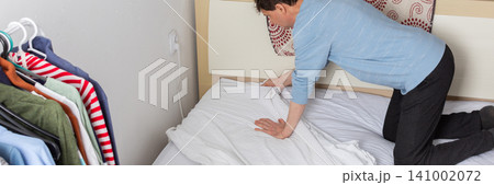 A 53-year-old man is changing the bed linen in his bedroom. He is pulling the fresh linen over the mattress while organizing the space for the day, banner A 53-year-old man is changing the bed linen in his bedroom. He is pulling the fresh linen over the mattress while organizing the space for the day, banner 141002072