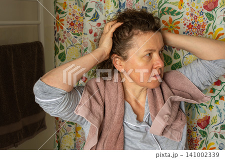 A 50-year-old woman prepares for her day in her bathroom. She is focused while removing a towel from her hair. The room is bright and colorful, reflecting a cheerful atmosphere A 50-year-old woman prepares for her day in her bathroom. She is focused while removing a towel from her hair. The room is bright and colorful, reflecting a cheerful atmosphere 141002339