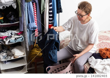 A person sits on the floor, sorting dirty clothes into a basket. The scene shows a cluttered laundry space with clothes piled on a chair and bed, indicating laundry day A person sits on the floor, sorting dirty clothes into a basket. The scene shows a cluttered laundry space with clothes piled on a chair and bed, indicating laundry day 141002403