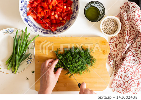 A person is chopping herbs on a wooden board while other fresh vegetables are arranged on a table. Bowls of diced tomatoes and salad dressing are also present 141002438