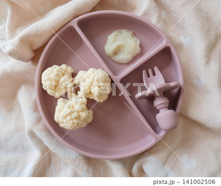 A baby is being fed with cauliflower florets using a plastic spoon on a baby plate from a top-down perspective on a table with pink and white tones, creating a neutral and clean setting. 141002506