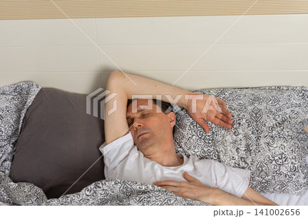 A 53-year-old man is asleep in his bedroom. He is lying on a bed covered with patterned bedding. His arm is stretched above his head. The room is quiet and inviting 141002656