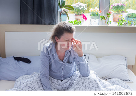 A tired 50-year-old woman sits on her bed, rubbing her eyes as she adjusts to the early morning light. Her cozy bedroom features plants and flowers visible outside the window A tired 50-year-old woman sits on her bed, rubbing her eyes as she adjusts to the early morning light. Her cozy bedroom features plants and flowers visible outside the window 141002672