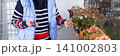 A woman tends to autumn flowers in pots on a balcony. She is 50 years old, wearing glasses and a sweater. The setting shows a typical urban view and seasonal flowers in bloom, banner 141002803