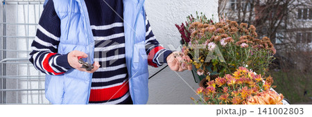 A woman tends to autumn flowers in pots on a balcony. She is 50 years old, wearing glasses and a sweater. The setting shows a typical urban view and seasonal flowers in bloom, banner A woman tends to autumn flowers in pots on a balcony. She is 50 years old, wearing glasses and a sweater. The setting shows a typical urban view and seasonal flowers in bloom, banner 141002803