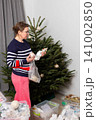 A person adds white ornaments to a Christmas tree. The tree is standing in a living room with gray curtains. The activity happens during the holiday season at home 141002850