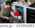 A man is putting dirty dishes into the dishwasher in his kitchen. He focuses on the task while several items rest in the appliance, ready for cleaning 141002883