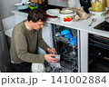 A man is at work in the kitchen, pouring salt into a dishwasher as he prepares to wash dirty dishes. He is focused on the task at hand while organizing the appliances 141002884