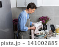 A man stands in the kitchen wiping clean dishes with a cloth. He wears an apron and focuses on his task. The kitchen has a sink and some dishes on the counter 141002885