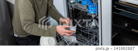 A man is at work in the kitchen, pouring salt into a dishwasher as he prepares to wash dirty dishes. He is focused on the task at hand while organizing the appliances, banner 141002887