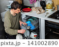 A man is putting dirty dishes into the dishwasher in his kitchen. He focuses on the task while several items rest in the appliance, ready for cleaning 141002890