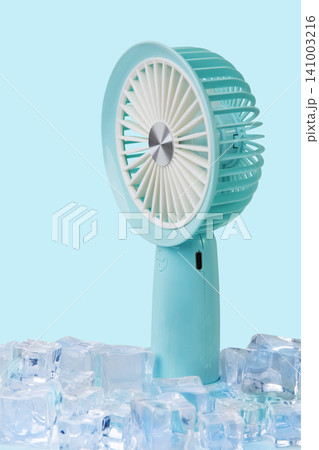 Blue portable mini fan standing among ice cubes on light blue background, extreme summer cooling concept 141003216